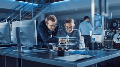 Dos ingenieros revisan un componente electrónico en un laboratorio de oficina técnica mientras un tercer profesional trabaja al fondo.