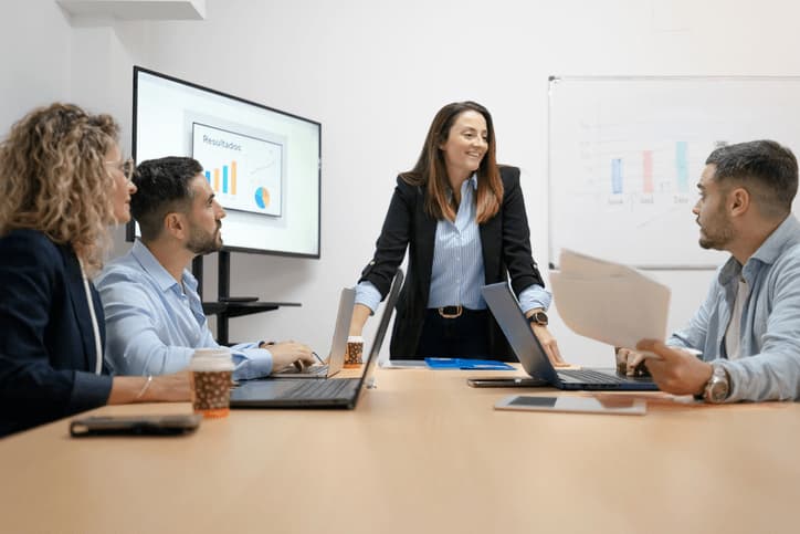 Dos directivos/as conversando en el exterior o en un pasillo acristalado de una planta industrial moderna, con vistas a la nave de fondo — uno de ellos mostrándole al otro una gráfica o tabla en una tablet, como si estuvieran revisando el dato de benchmarking en ese momento. Aporta el punto humano (conversación, decisión compartida) y el contexto industrial serio de fondo, sin repetir el despacho cerrado del artículo anterior.